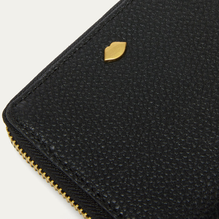 Close-up of a black pebbled leather zip-around wallet with a small gold-tone lip-shaped stud and gold zipper on a light neutral background.