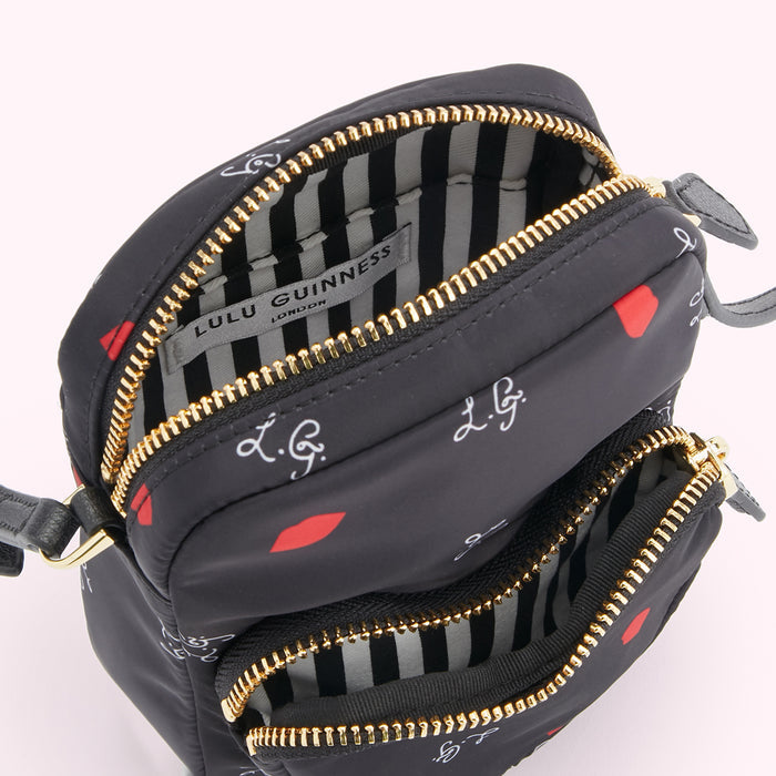 Close-up view of a black Lulu Guinness crossbody bag with red lips and the designer's initials printed on it. The bag is open, showing its striped interior lining and gold zippers.