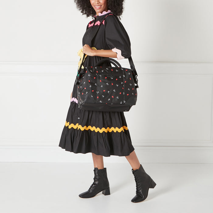 A woman in a black dress and black boots carries a black duffel bag with a red lips and white bird print.