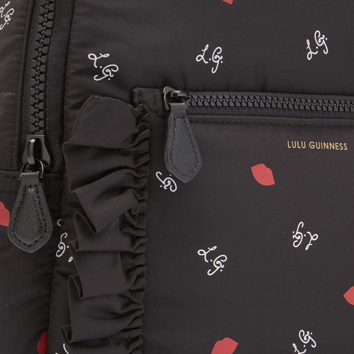 Close-up of a black Lulu Guinness backpack with a ruffled detail, featuring a repeating pattern of red lips and the brand's logo.