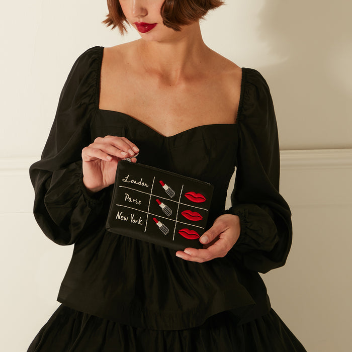 A black makeup bag with embroidered lips and lipsticks, and the words "London," "Paris," and "New York" on it, is held by a person wearing a black top.
