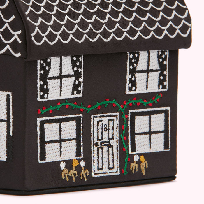 Close-up of a black, house-shaped box with white embroidered details, including windows, a door, roof shingles, and a garland of lights.
