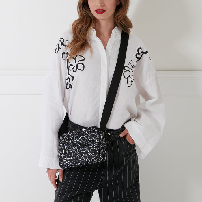 Woman wearing a white shirt with black floral print and black pinstripe pants, carrying a black crossbody bag with white embellishments. The background is white.