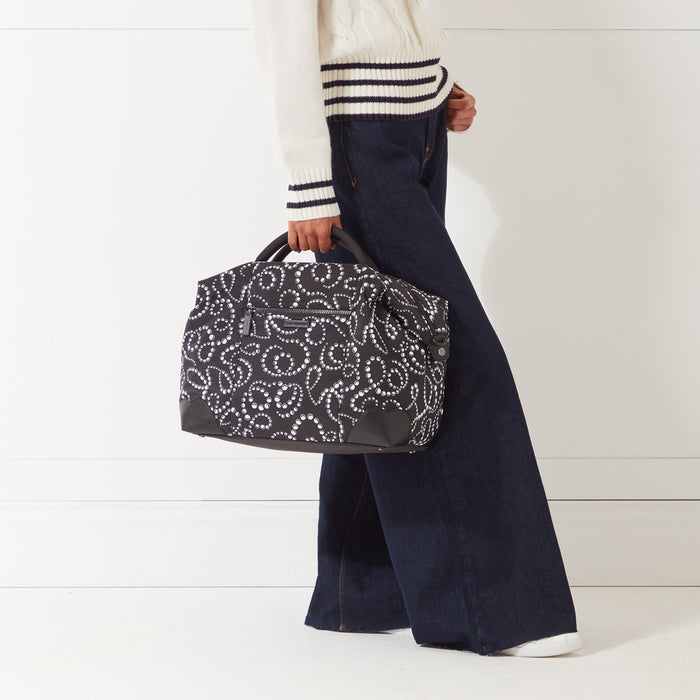 A woman in dark jeans and a cream sweater carries a dark-coloured travel bag with a white pearl pattern. The background is white.