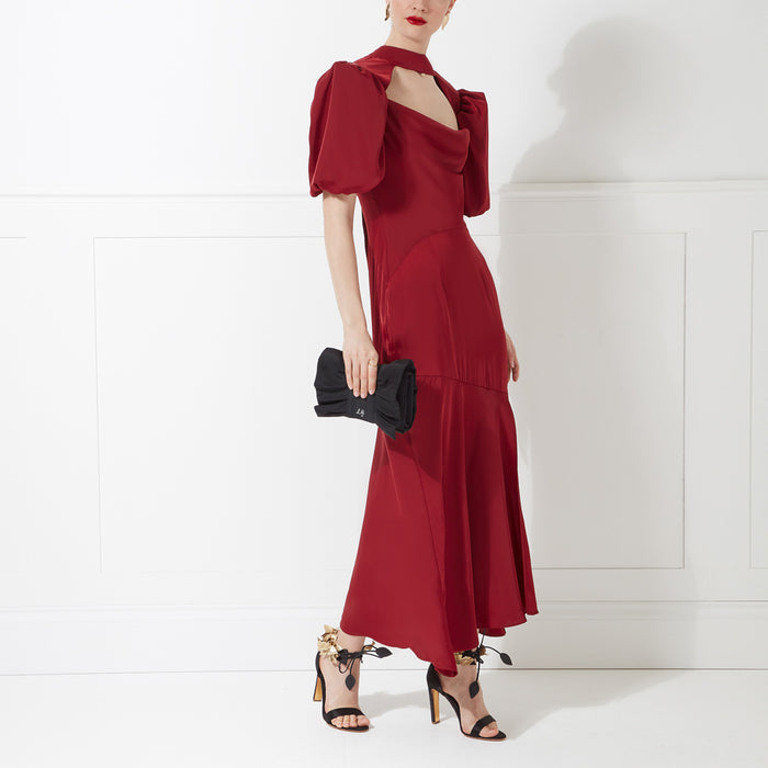 Woman in a red midi dress with puff sleeves, holding a black clutch. She is wearing black and gold heels. The background is white.