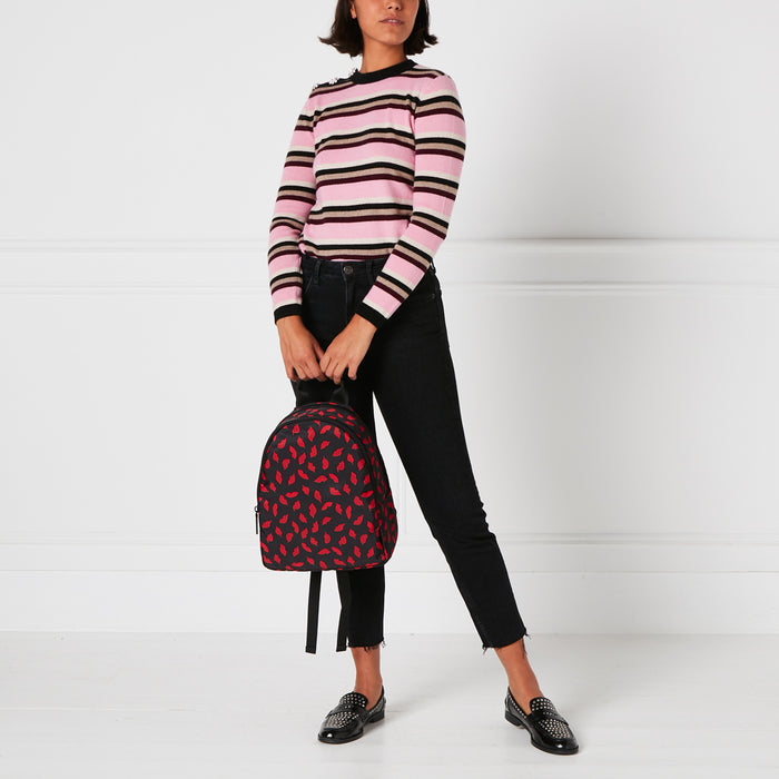 A person with short brown hair wearing a pink and brown striped sweater and black pants is standing in front of a white background. The person is holding a black backpack with a red lips pattern.