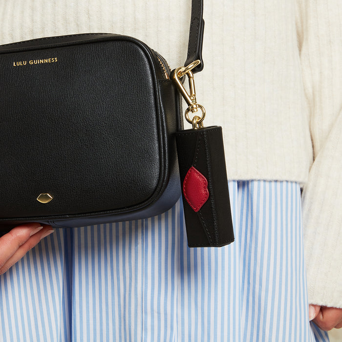 A black and red leather lip key charm clipped onto a black bag and held by a person wearing a white sweater and blue striped shirt.