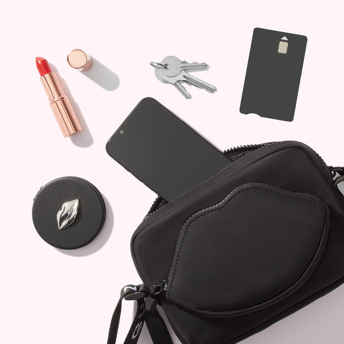 A black crossbody bag with a lip-shaped pocket lies open, displaying its contents against a white background: a smartphone, lipstick, compact mirror, keys, and a SIM card.