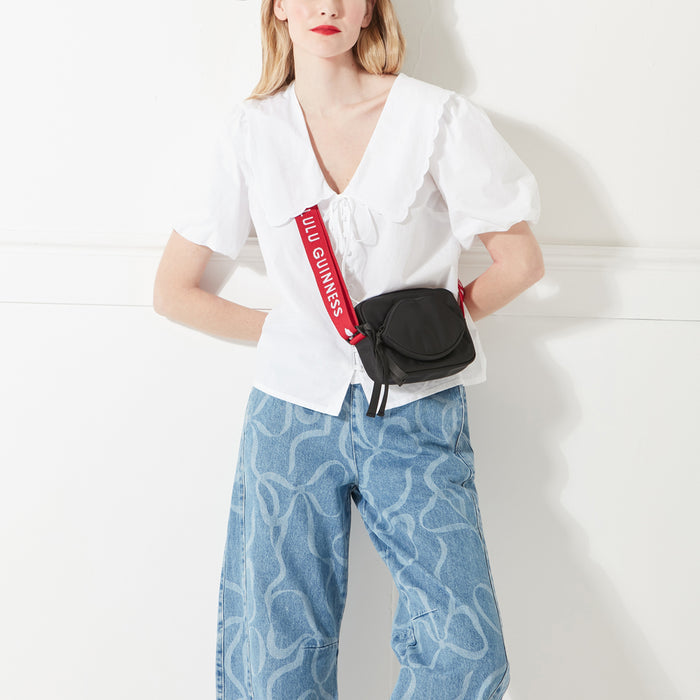 A blonde model standing against a white background wearing a white top, blue denim jeans and a black nylon crossbody bag with a lip pocket and red 'Lulu Guinness' branded strap.