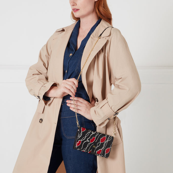 Woman in beige trench coat and dark wash jeans holding a black crossbody bag with red lips and gold chain.