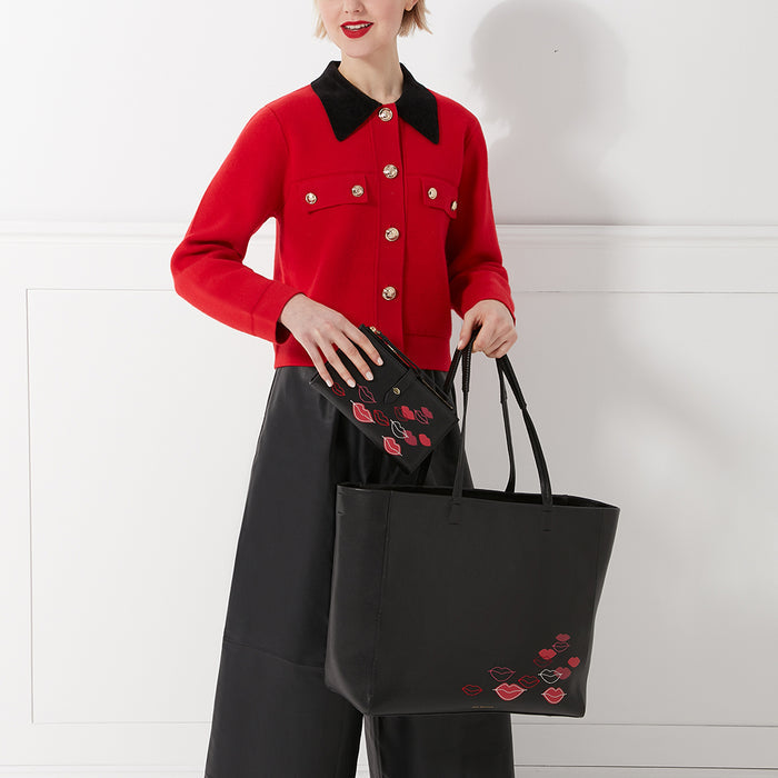 A woman in a red cardigan holds a black tote bag and a smaller black wallet, both decorated with red and pink lips. The background is white.