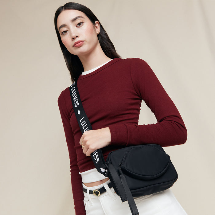 Woman holding a black crossbody bag with 'Lulu Guinness' branding on the strap against a beige background