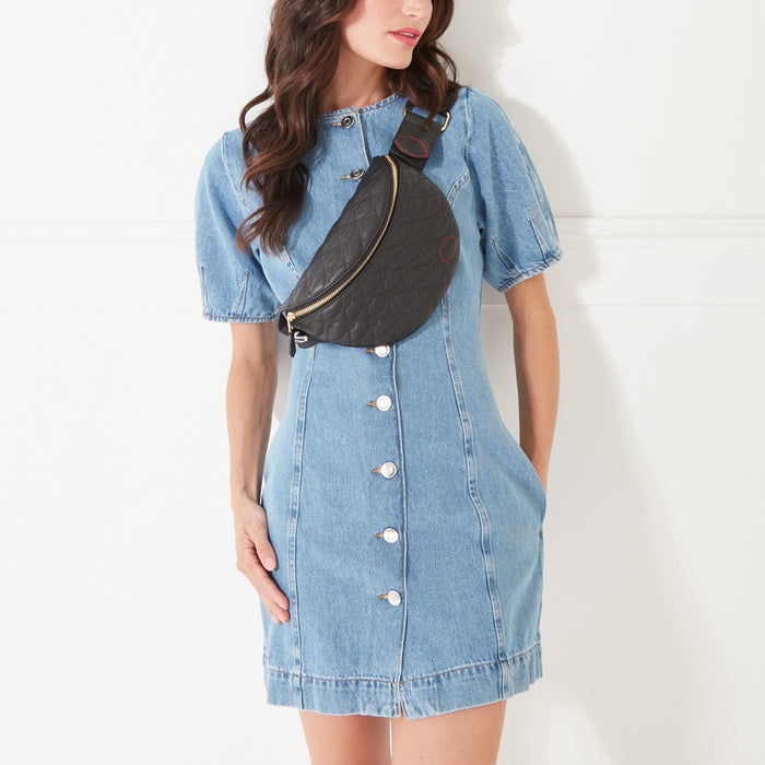 A model with dark hair wearing a black leather belt bag with lip shaped quilting across her body in a denim dress.