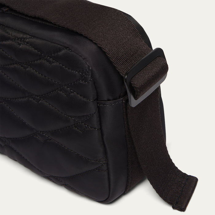 Close up image of a black quilted crossbody bag with a woven belted strap on a light gray background