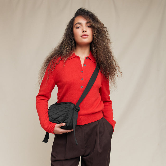 Person wearing a red sweater holding a black quilted crossbody bag against a beige background