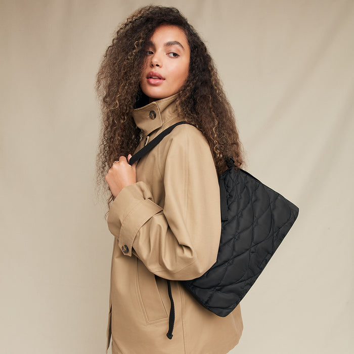 Woman wearing a beige coat with a black quilted bag over her shoulder against a beige background
