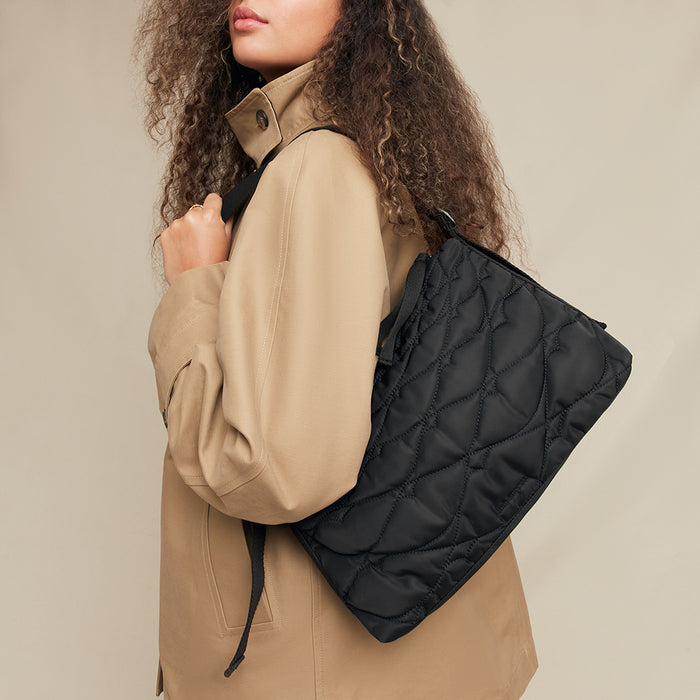 Person wearing a beige jacket with a black quilted bag over their shoulder against a neutral background