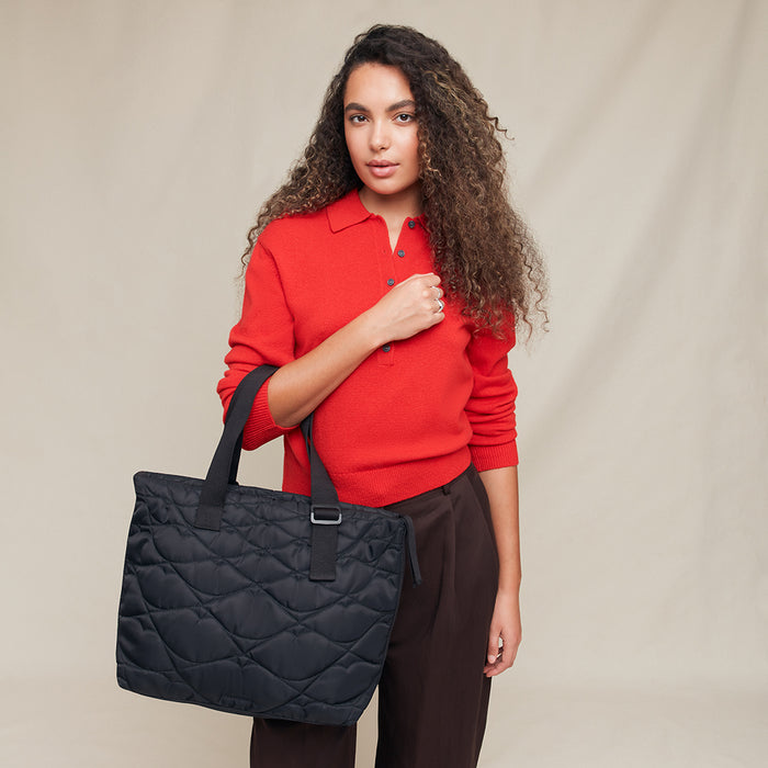Woman in a red sweater holding a black quilted tote bag against a beige background