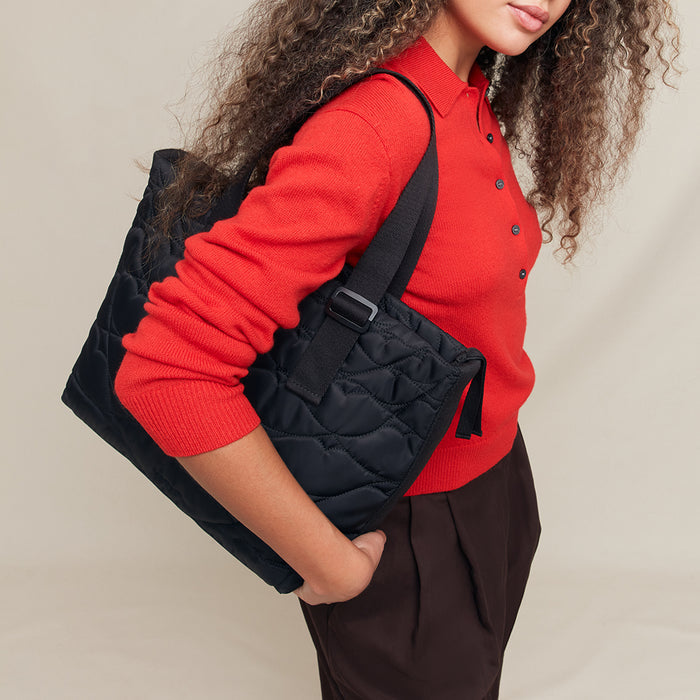 Person wearing a red sweater and holding a black quilted tote bag against a neutral background