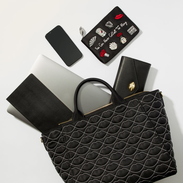 A black quilted tote bag with a gold zipper sits on a white surface with a silver laptop, a black phone, a black wallet and a black embroidered pouch inside.
