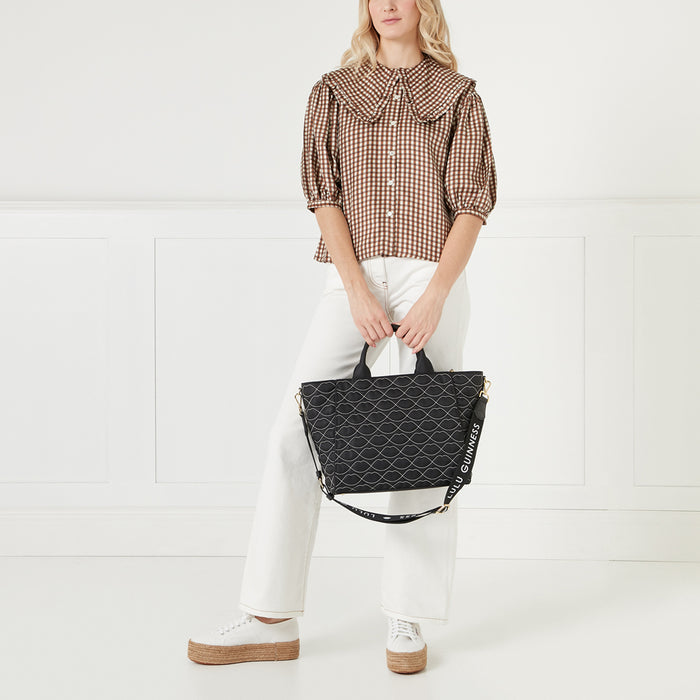A woman in a brown gingham shirt and white pants holds a black Lulu Guinness tote bag with a lip print. The bag has a black strap with the brand name written on it. The background is white.
