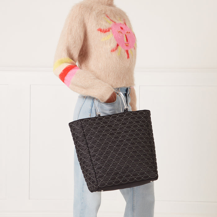 Woman in light beige fuzzy sweater and light blue jeans holding a large black quilted tote bag with clear handles. The bag features a repeating lip print. The background is white.
