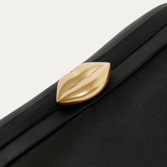 Close-up of a black satin clutch bag with a gold lip clasp on a light gray background