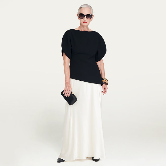 Woman wearing a black top and white skirt with sunglasses holding a satin clutch bag on a plain background