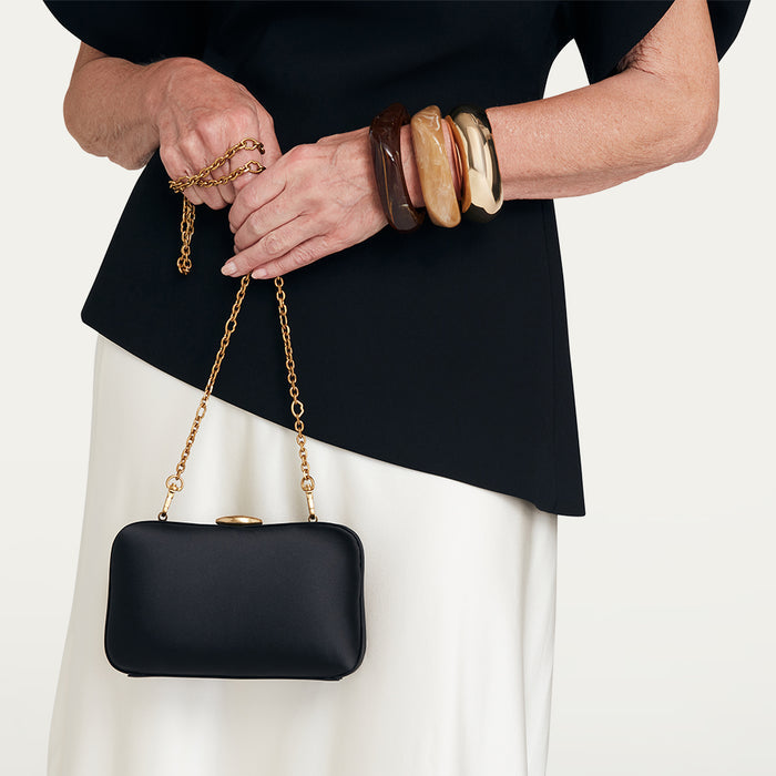 Person holding a black satin clutch with a gold chain strap against a plain background
