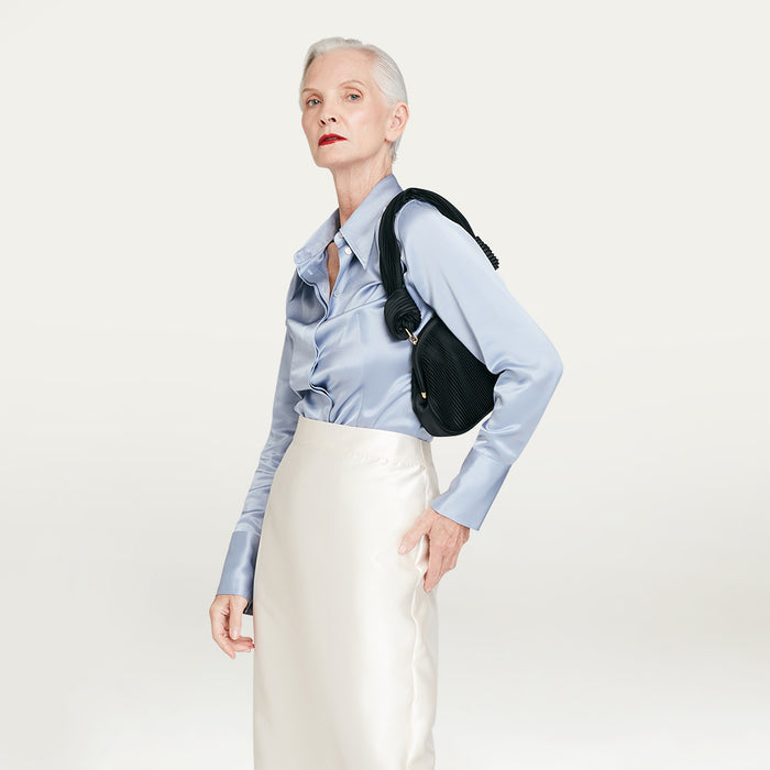 Woman wearing a light blue shirt and white skirt with a black bag on a plain background