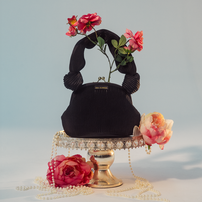 Black satin pilisse handbag on a stand with flowers and pearls.