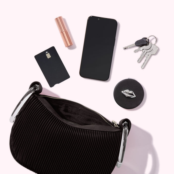 A black handbag sits open, displaying its pleated interior. Surrounding it are a smartphone, lipstick, credit card, keys, and a compact mirror with a lip design. The items are arranged on a light pink background.