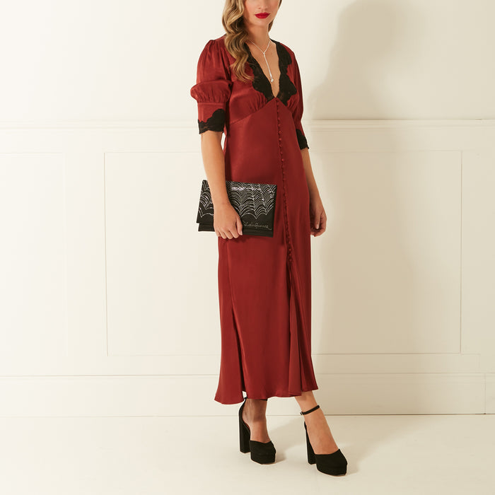Woman in a rust-coloured midi dress and black heels holds a black clutch.