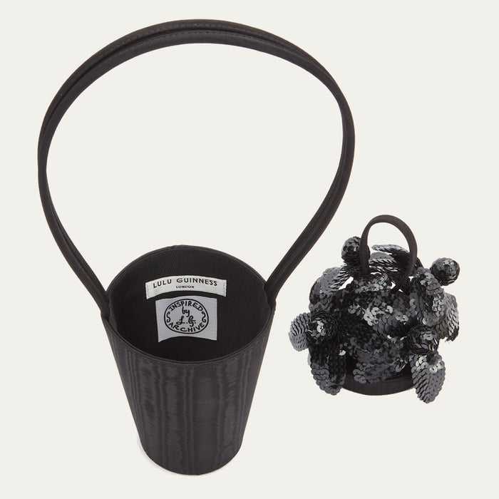 Black satin moire bucket bag with top looped handle showing inside bag revealing the branded 'Lulu Guinness London' and 'Inspired by the Archive' labels. Sitting next to the bag lid covered in black sequin flowers.