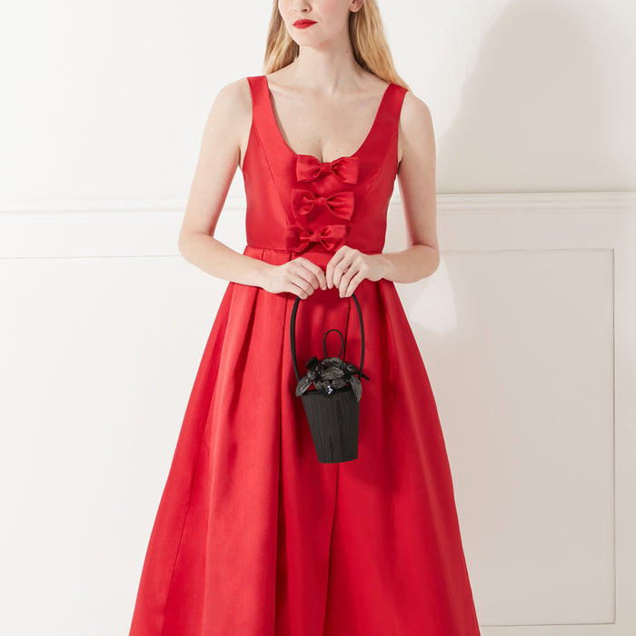 Model in red satin dress with bow details holding a black satin handbag with black sequin lily flowers on the top.
