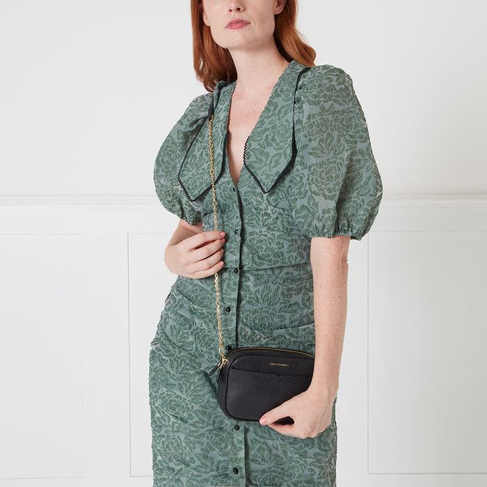 Woman in sage green floral dress holding a black crossbody bag with gold chain strap.