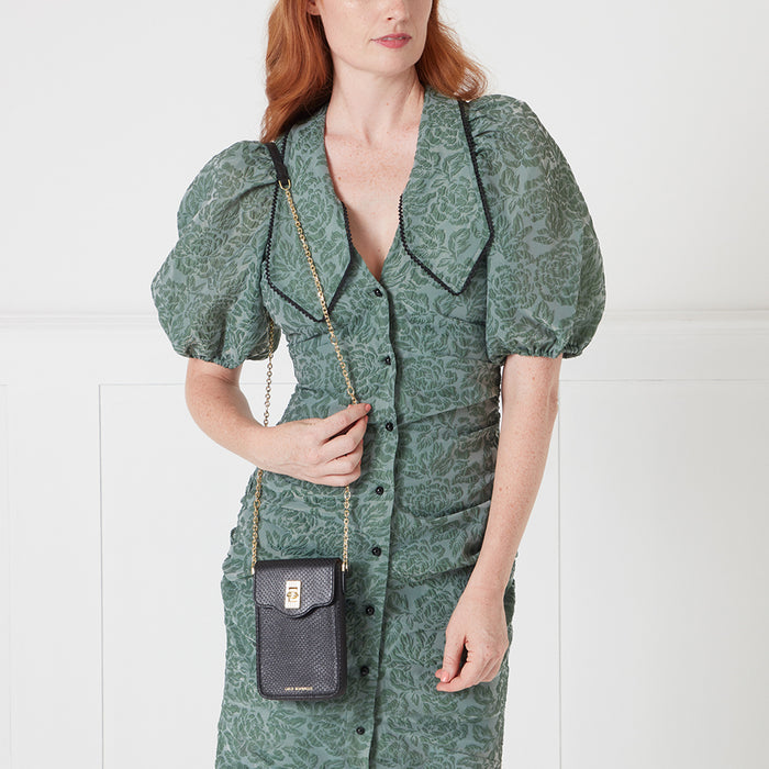 Woman in sage green floral dress holding black crossbody phone bag with gold chain strap.