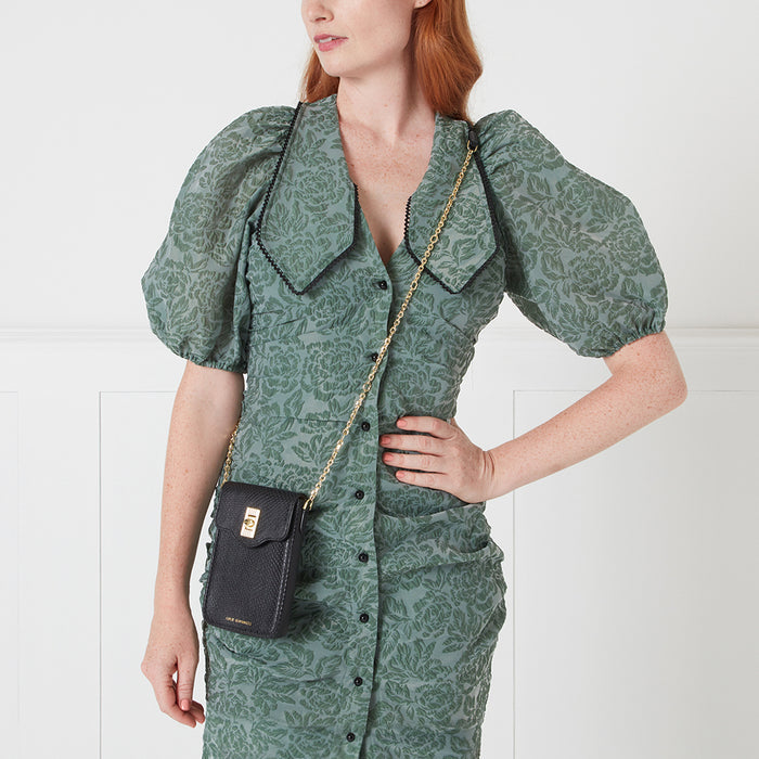 A woman in a sage green floral dress carries a small black crossbody bag with a gold chain strap.
