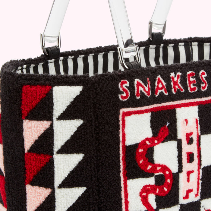 Close-up of a black tote bag with colourful embroidered geometric patterns and a snake on a checkerboard. The words "SNAKES" are embroidered above the checkerboard. The bag has clear acrylic handles.