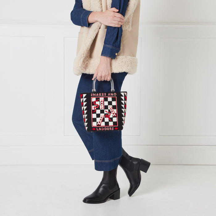A woman in a denim shirt and cream shearling vest holds a black and white Snakes and Ladders-themed tote bag with a clear acrylic handle.