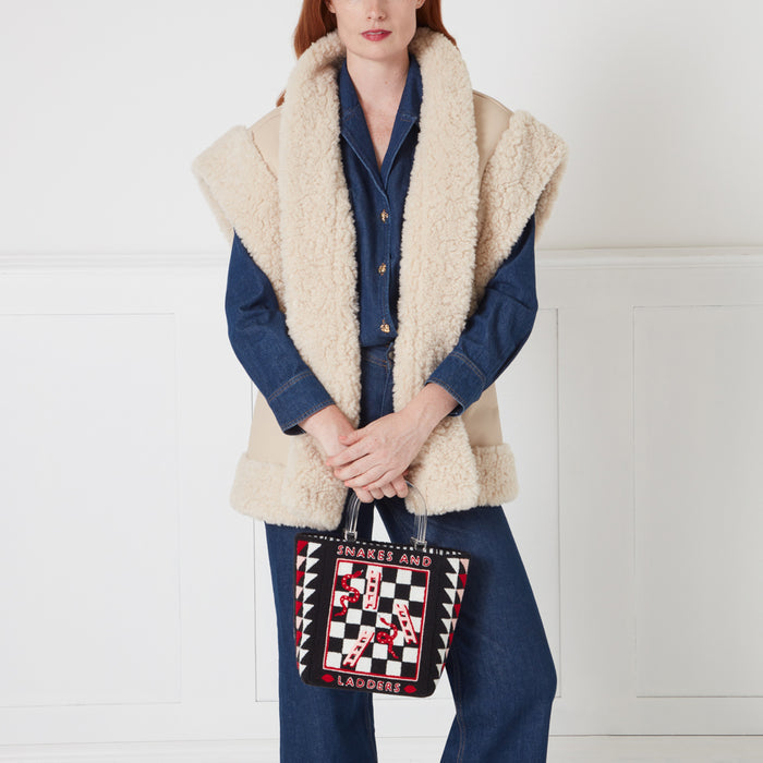 Woman in denim shirt and cream shearling vest holding a black and white snakes and ladders tote bag.