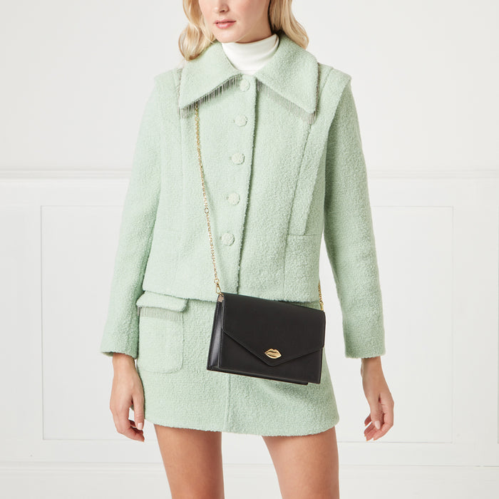 A woman in a light green jacket holds a black crossbody bag with a gold lip-shaped detail.
