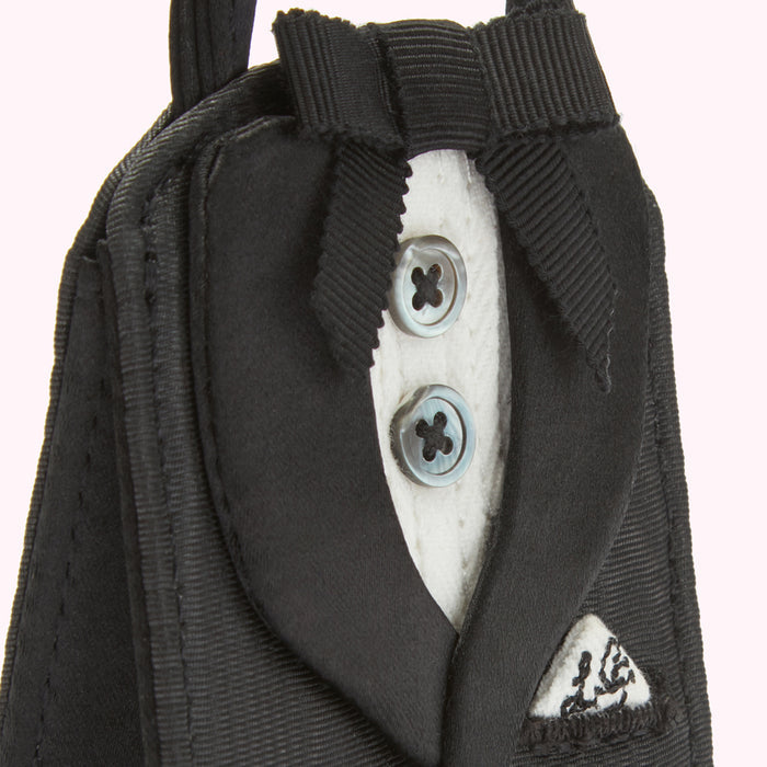 Close-up of a tuxedo-shaped ornament with black fabric, white shirt detail, two grey buttons, and a black bow tie. The background is white.