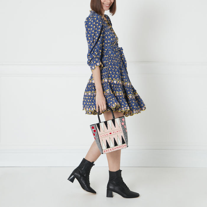 A woman in a blue floral dress holds a backgammon-themed tote bag. The bag is cream with black and pink details. She is wearing black ankle boots. The background is white.