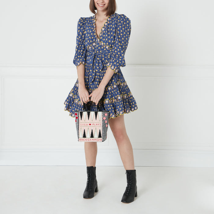 A woman in a blue floral dress holds a black and white backgammon-themed tote bag against a white background.
