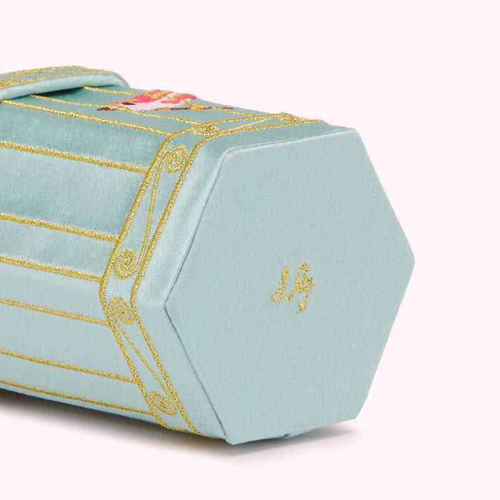 Close-up of a pale teal hexagonal box with gold embroidery depicting a carousel scene and the letter "LG" embroidered on the bottom.