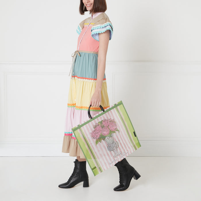 A woman in a colorful tiered dress carries a large tote bag featuring a pink rose illustration against a white background.