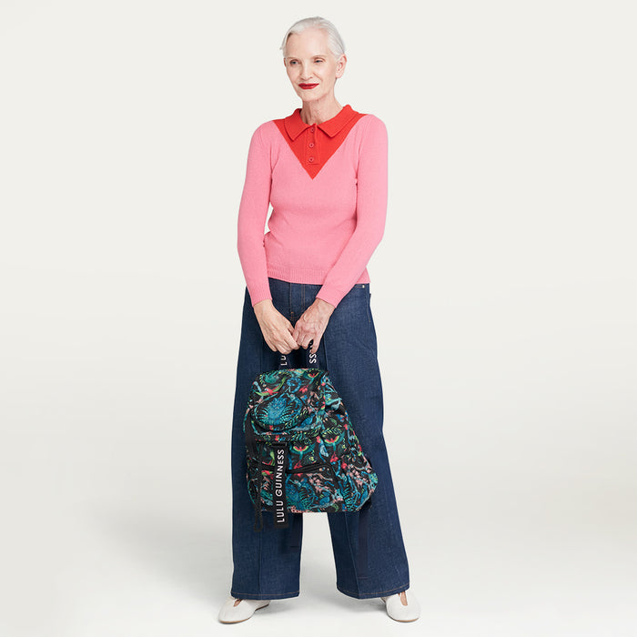 Woman wearing a pink sweater with a red collar and blue jeans, holding a floral Lulu Guinness x Emma Shipley backpack on a light gray background
