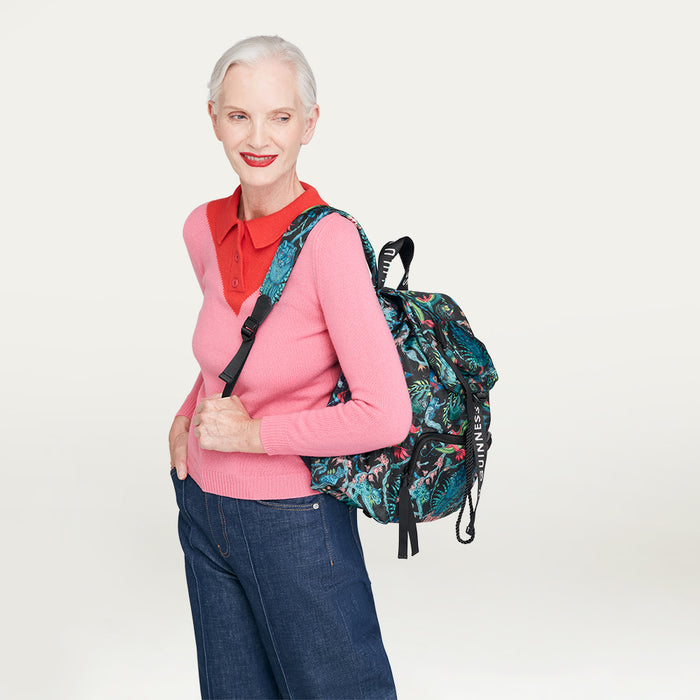 Woman wearing a pink sweater and blue jeans with a floral printed Lulu Guinness x Emma Shipley backpack on a light gray background