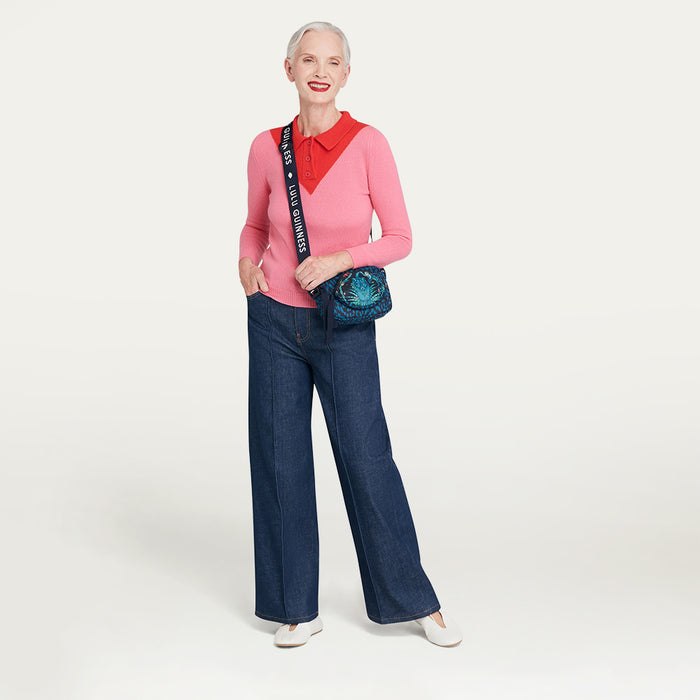 Woman wearing a pink sweater with a red collar, blue jeans, and wearing a printed Lulu Guinness x Emma Shipley crossbody bag on a white background